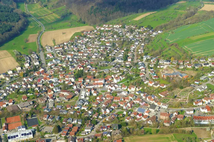 Luftbild der Ortsmitte von Wiesenbach von Süden nach Norden. Das Rathaus ist der hellrote Bau mit Türmchen …